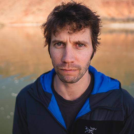 A man with messy brown hair and a serious expression, wearing a dark and blue jacket, against a blurred warm background.