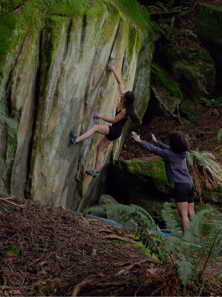 Two women bouldering outdoors, one climbing a mossy rock and the other spotting.
