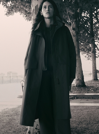 Monochromatic photo of a woman with long hair wearing a long coat, leaning against a tree and looking up.