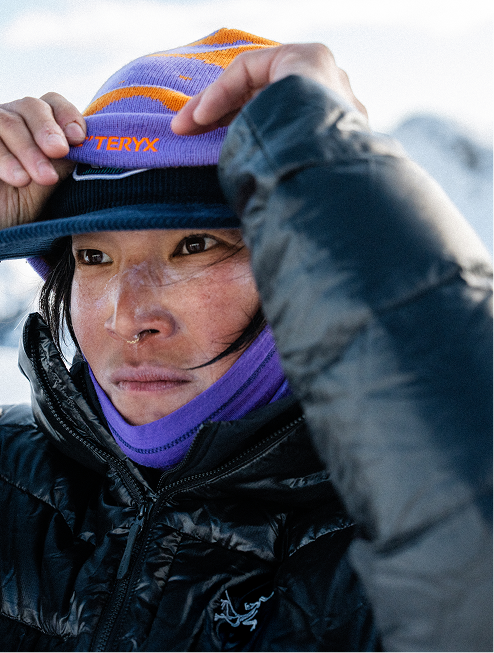 A person adjusts their purple and orange Arc'teryx beanie, wearing a black puffy jacket and sun protection on their face in a snowy environment.