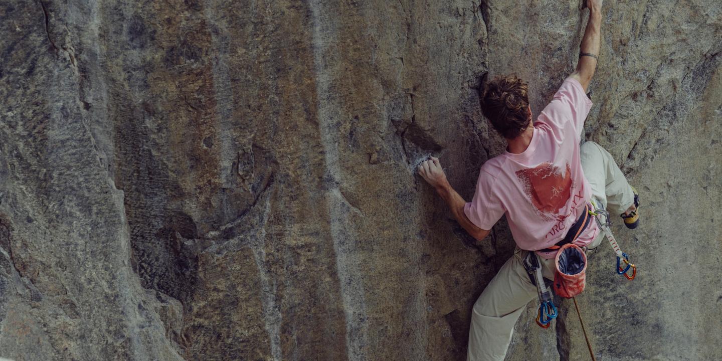 A man in a pink t-shirt rock climbing on a grey rock wall, seen from behind.