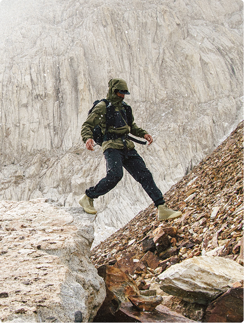 A person in olive green hiking gear steps over rocks on a snow-dusted mountain.