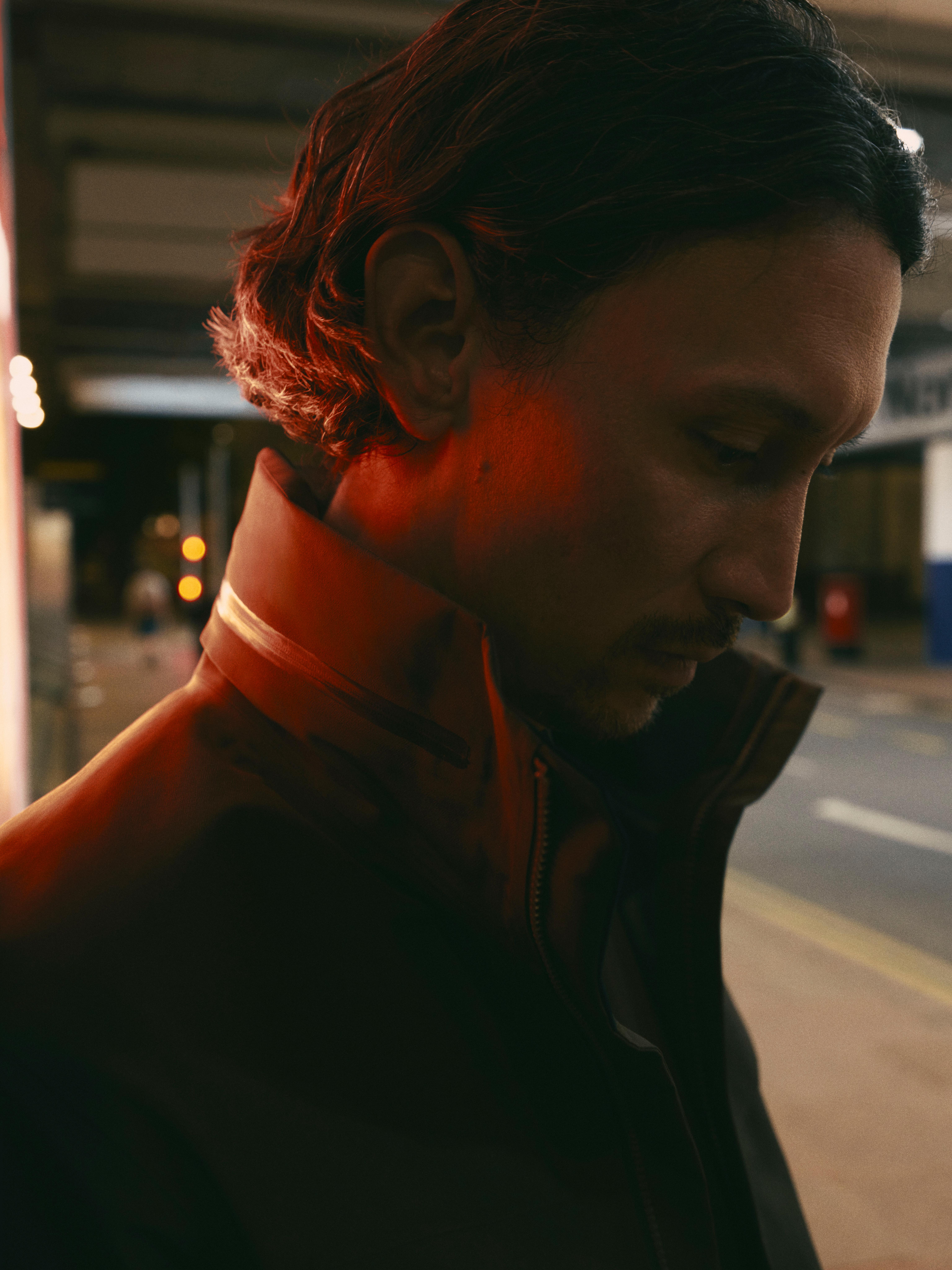 A man's profile, head bowed, with dark hair and a dark jacket, illuminated by a red glow from behind.