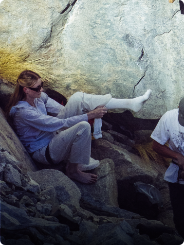 A woman in sunglasses and a harness sits on rocks, putting on a sock.