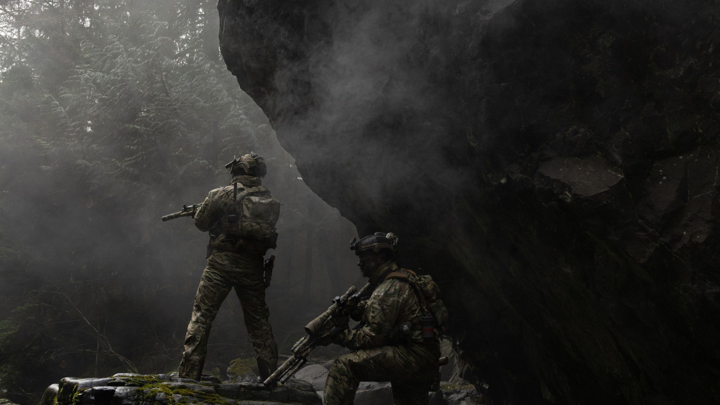 Two armed soldiers in tactical gear in a misty, rugged landscape.