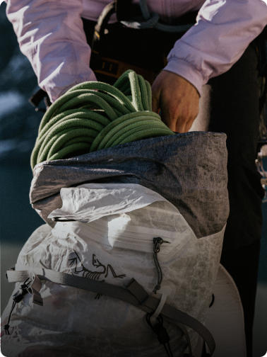 A person's hands packing a green climbing rope into a white Arc'teryx backpack.