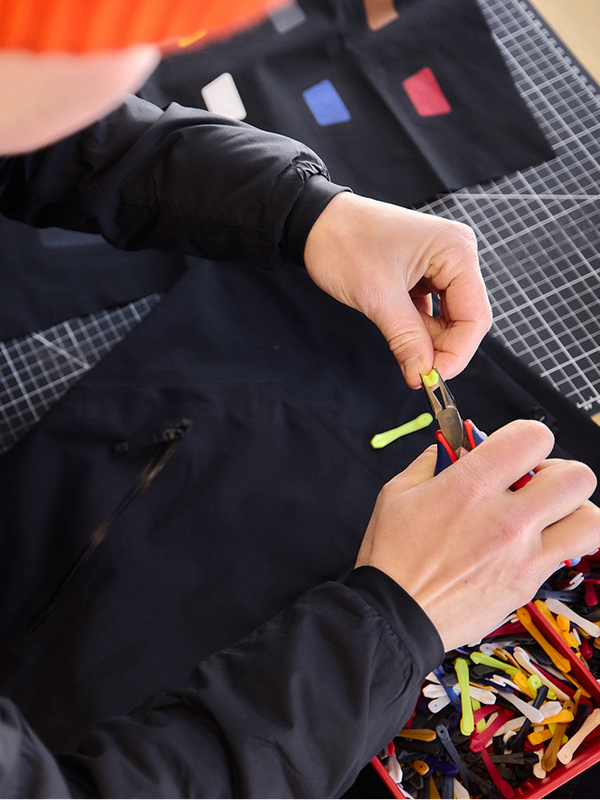 A person attaches a yellow zipper pull to a black jacket using pliers.