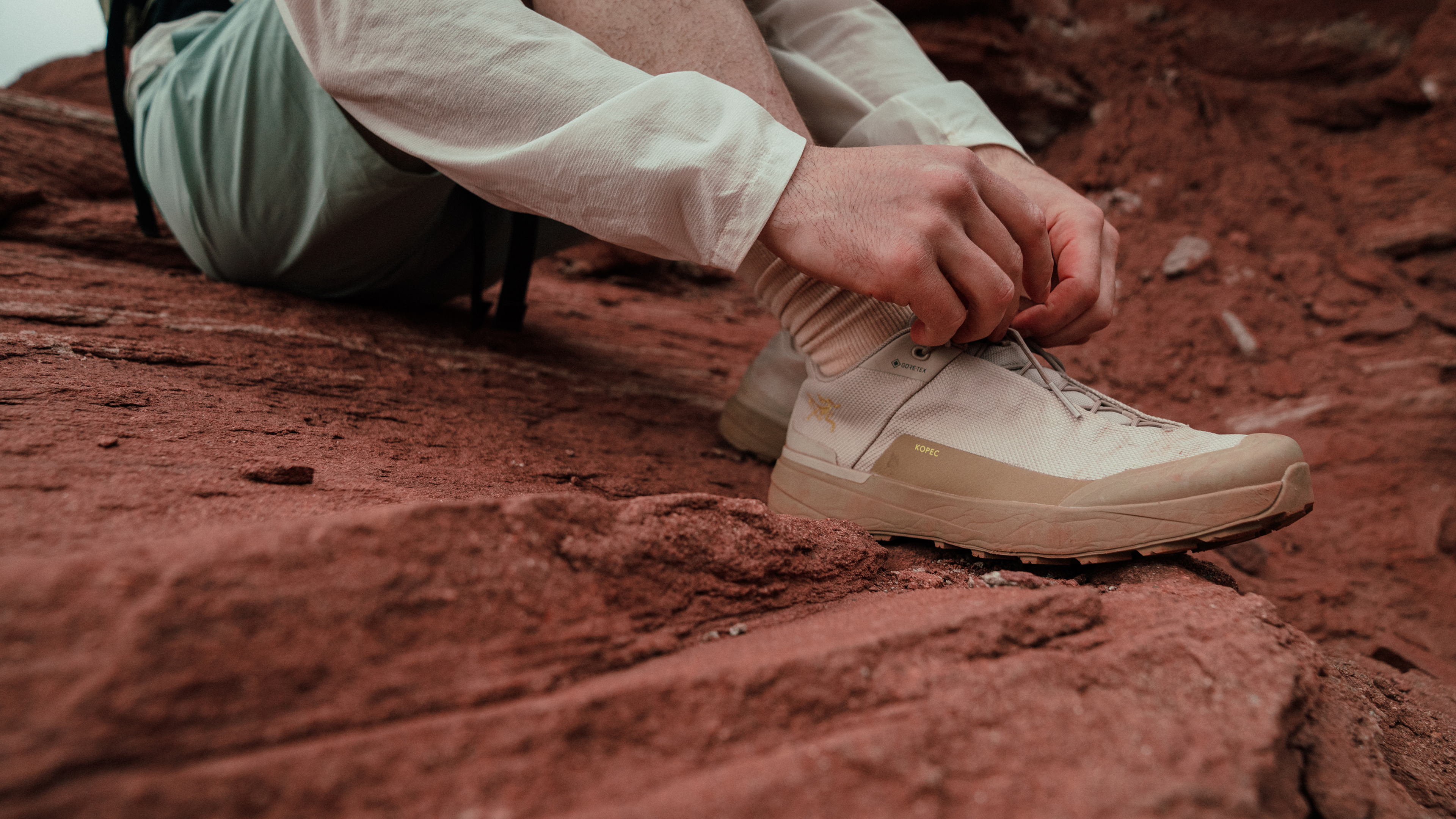 Person tying a tan Arc'teryx hiking shoe on red rocks.