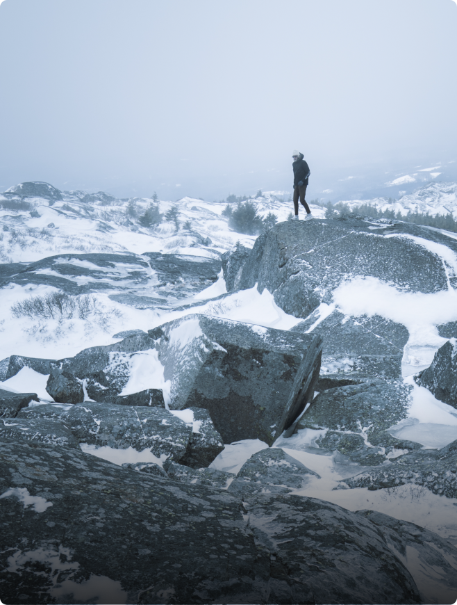 Person standing on a snow-covered rock in a hazy winter mountain landscape.