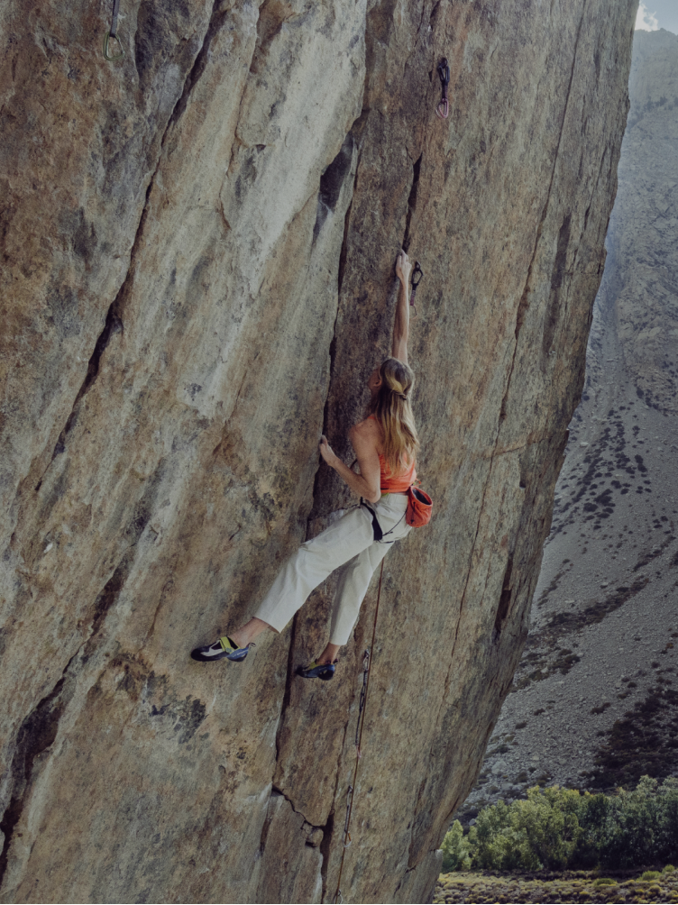 A woman in an orange top and light pants rock climbs a steep rock face, her right arm extended high for a handhold.