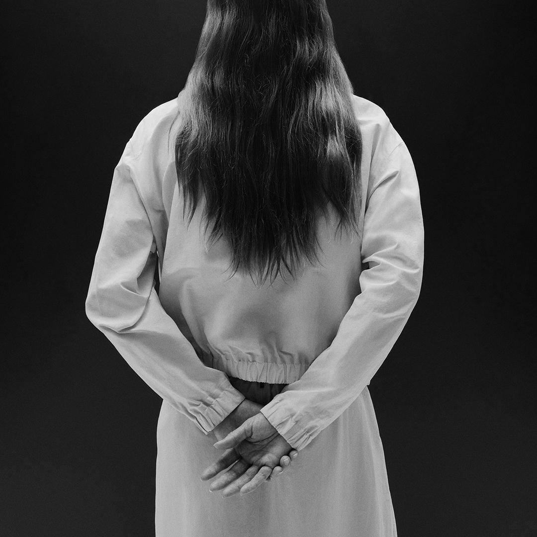 Black and white photo of a person from behind with long hair and hands clasped behind their back.