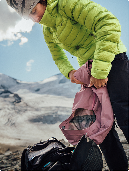 A hiker in a green puffer jacket and Arc'teryx beanie holds a pink jacket over a backpack on a snowy mountain.