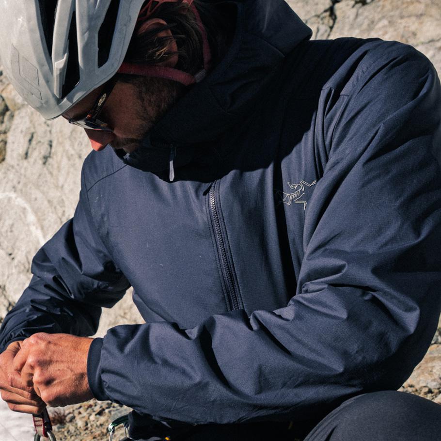 A person in a dark blue Arc'teryx hooded jacket, helmet, and sunglasses, adjusting climbing gear on a rocky surface.