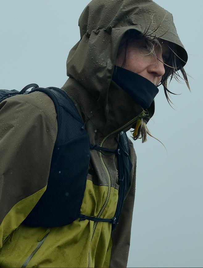 Person in a wet, hooded olive green rain jacket and running vest, with windblown hair.