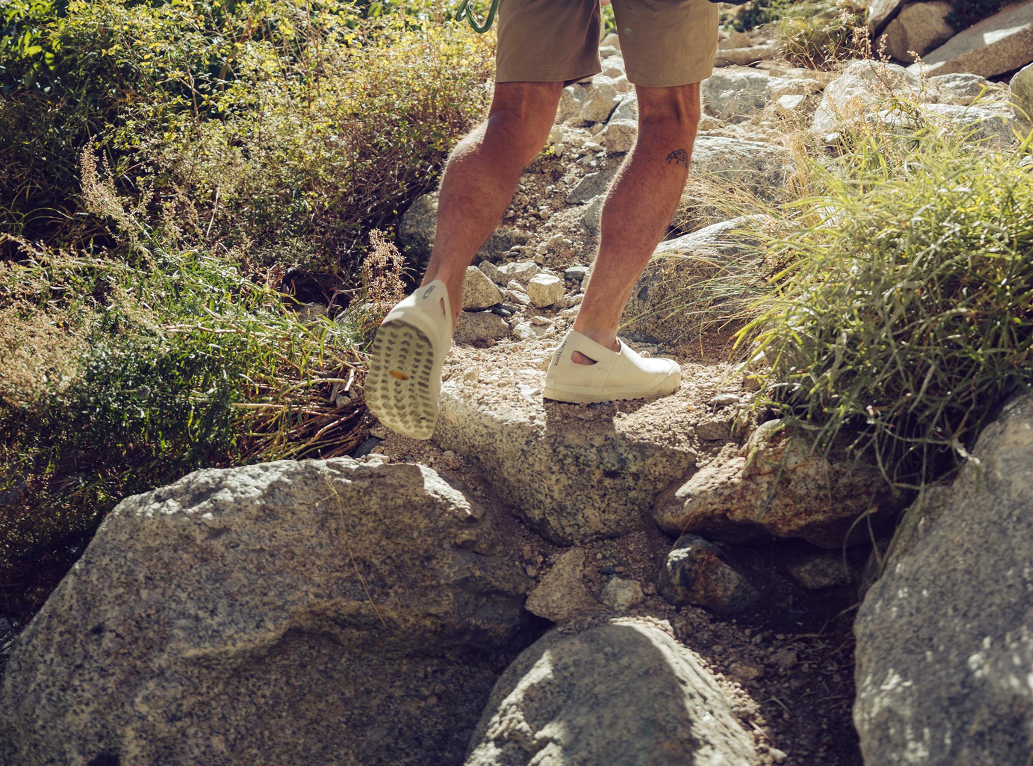 A person wearing shorts and light slip-on shoes walks on a rocky trail.