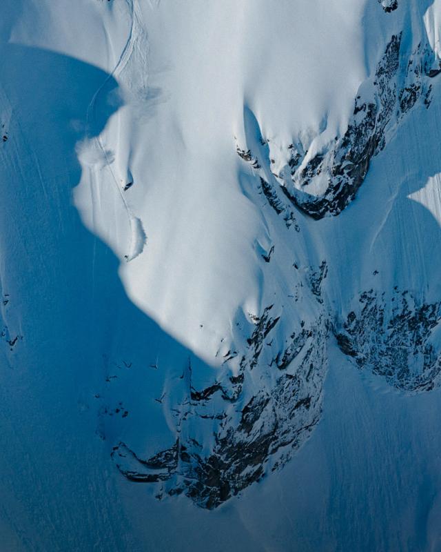 Aerial view of a skier descending a steep, snow-covered mountain slope with prominent shadows.