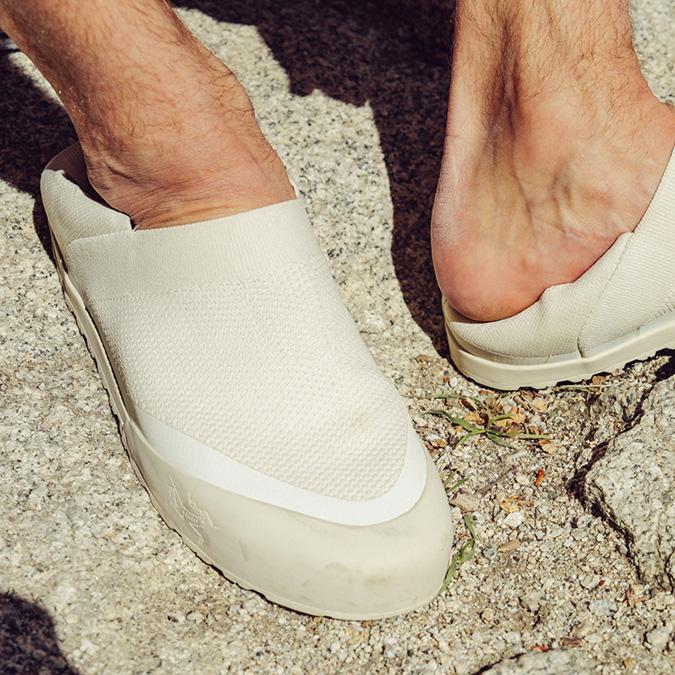 A person's feet in light-colored slip-on shoes on a rocky surface.