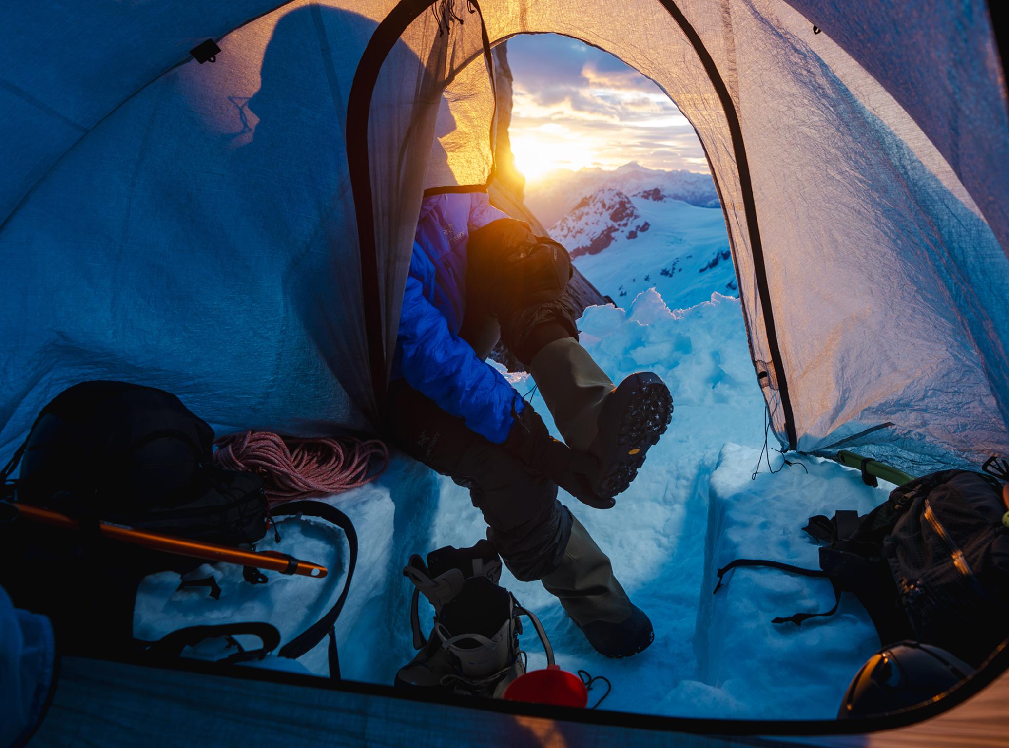 Person's legs extend from a tent into a snowy mountain range at sunset.