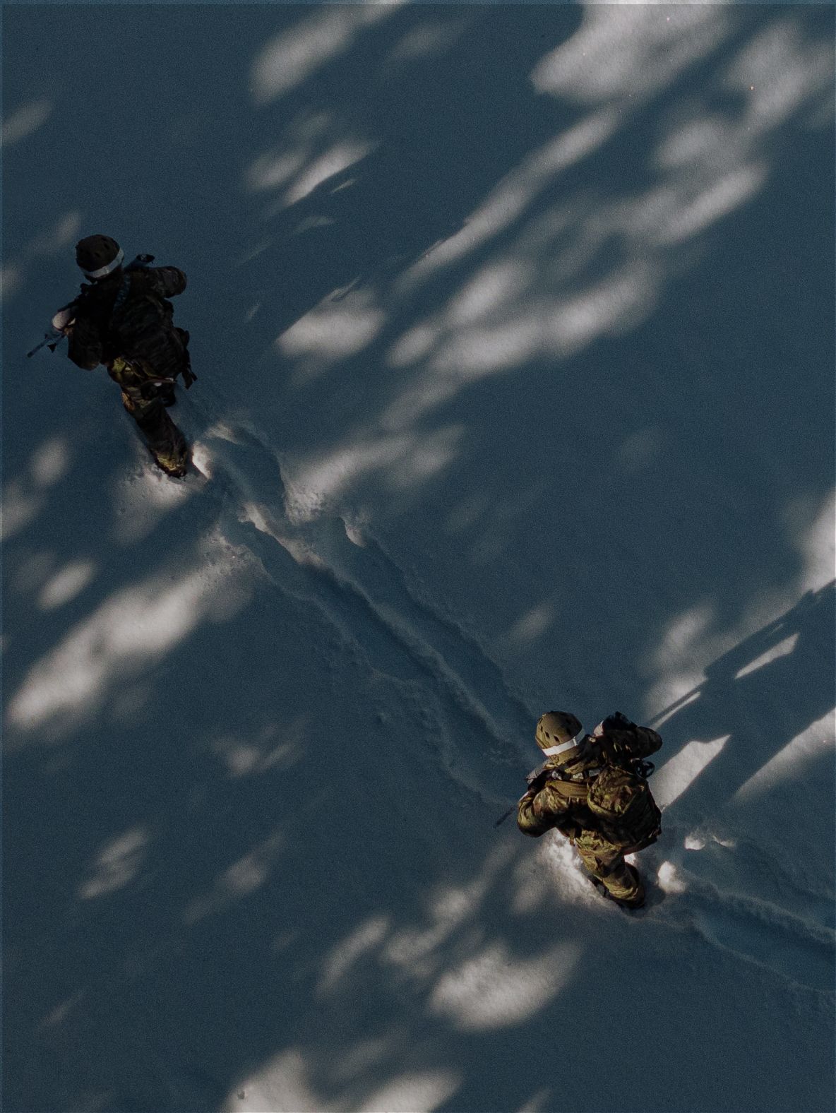 Overhead view of two soldiers walking through snow with dappled tree shadows.