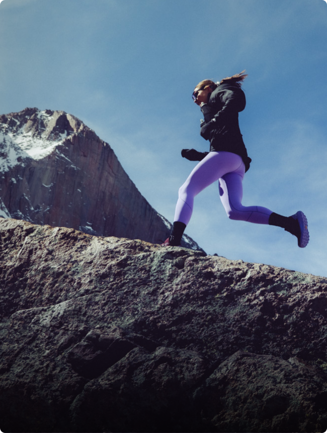 Woman in purple leggings leaping over rocky terrain with a snow-capped mountain in the background.