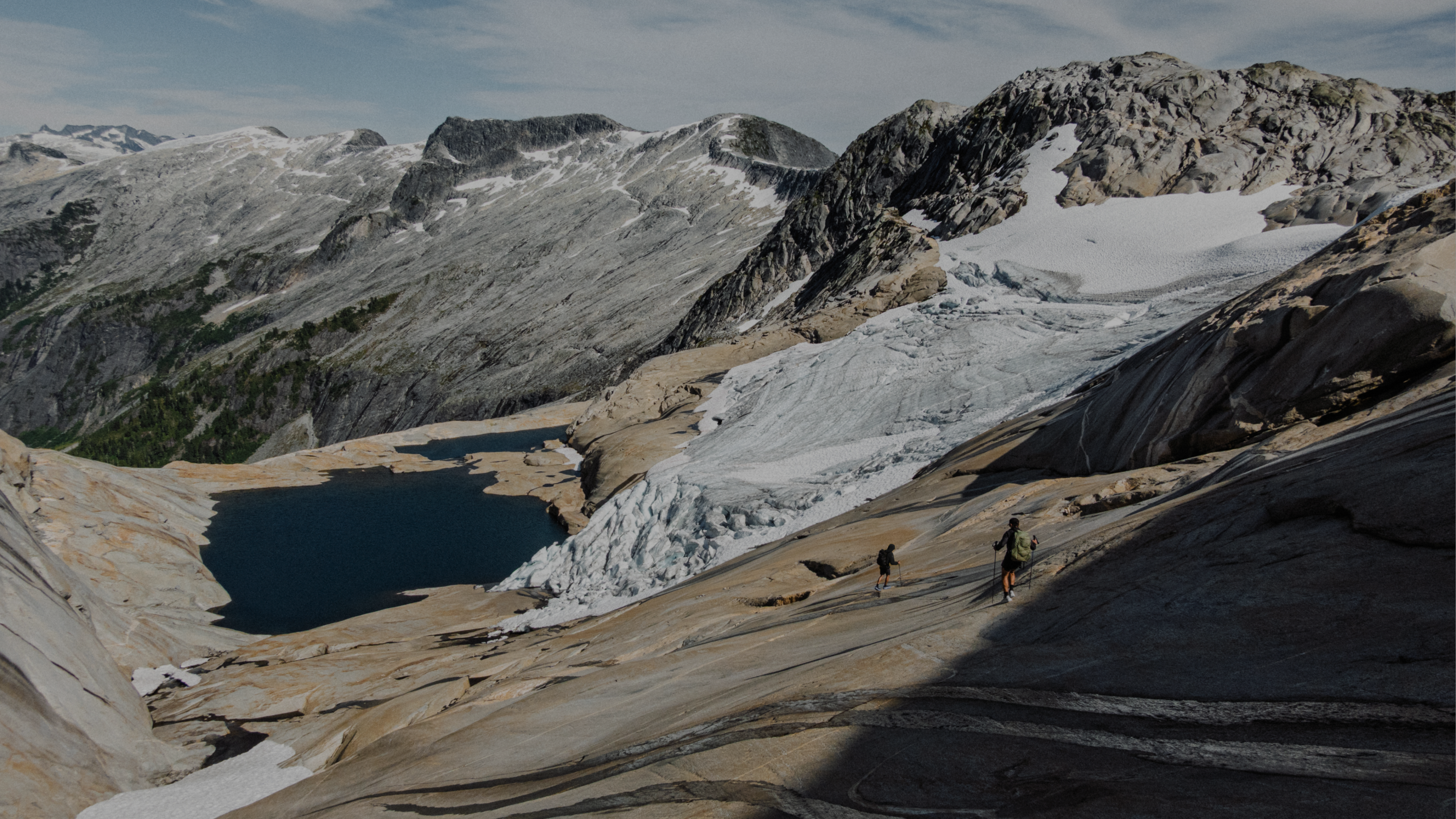 Two hikers on a rocky mountain slope with a glacier and a lake.