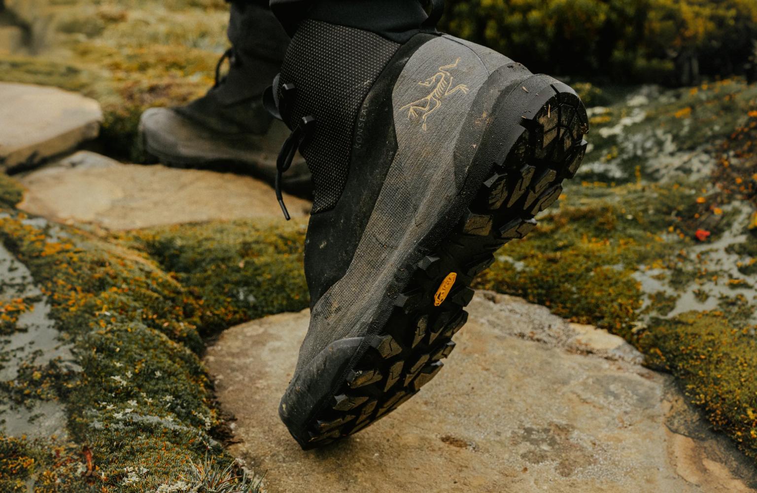 A dark Arc'teryx hiking boot with a Vibram sole steps onto a mossy stone path.