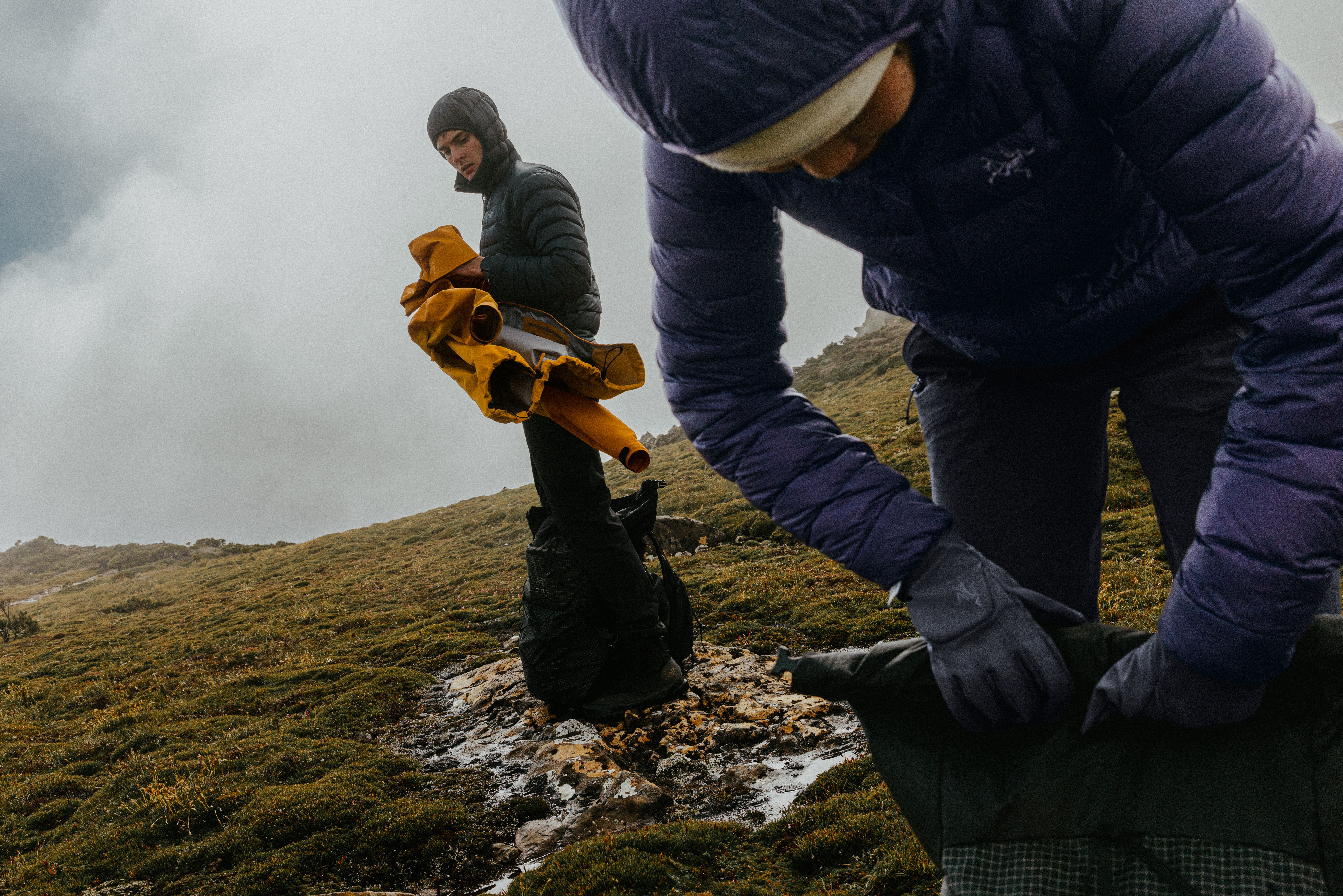 Two people in winter gear on a misty mountain trail, one holding a yellow jacket, the other packing a bag.