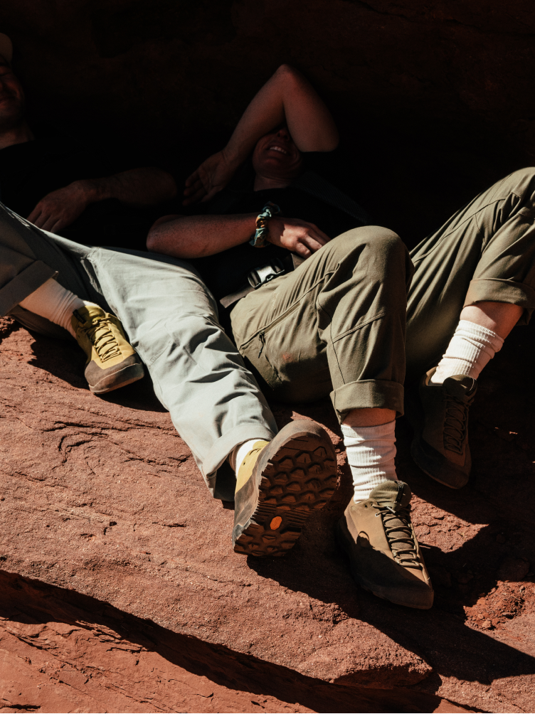 Two people wearing hiking pants and shoes sit on red rock.