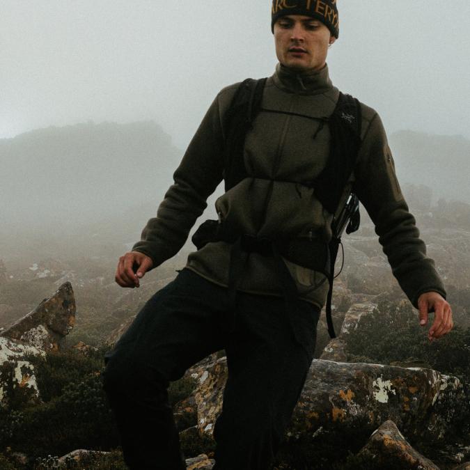 A man in an Arc'teryx beanie and hiking gear navigates a foggy, rocky mountain path.