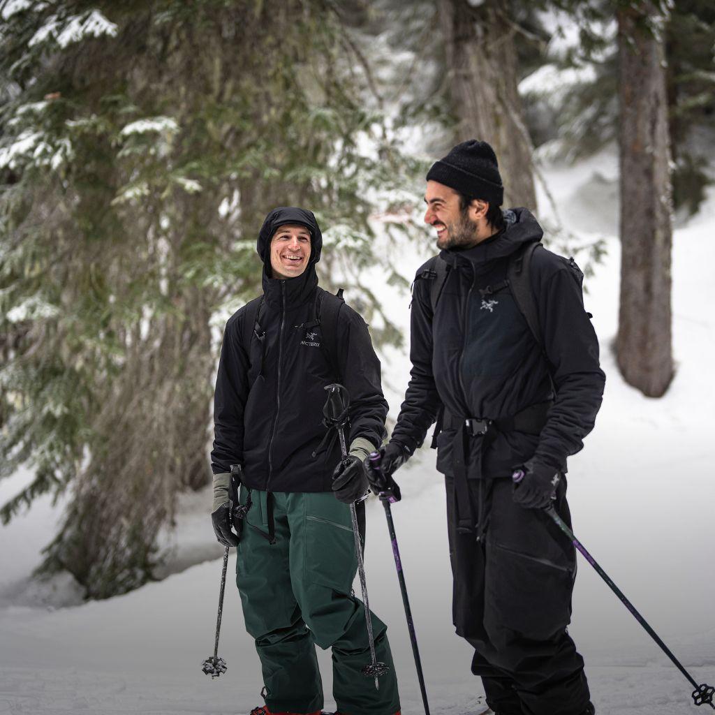 Two smiling men in ski gear stand in a snowy forest.