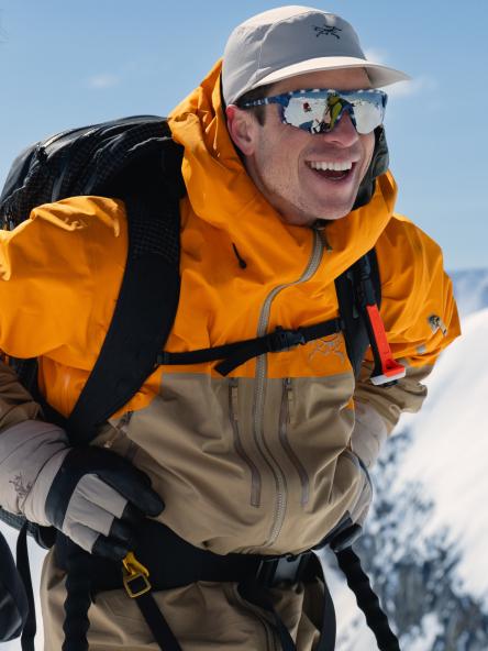 A smiling man in an orange and tan winter jacket, grey cap, and mirrored sunglasses with a backpack in a snowy setting.