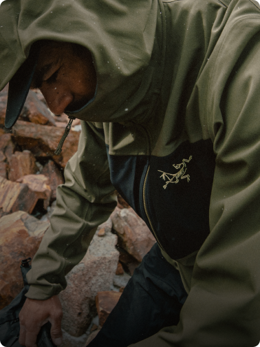 A person in an olive green and black Arc'teryx jacket, looking down amidst rocky terrain.