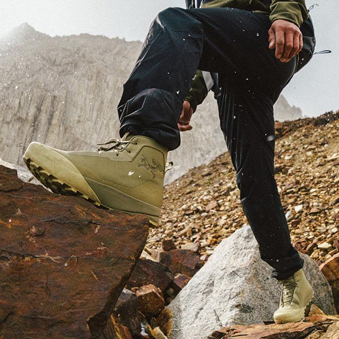Hiker in olive green boots stepping onto a large rock in a rocky, snowy mountain environment.