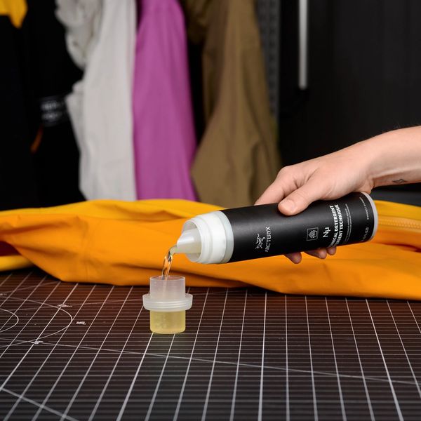 A hand pours Arc'teryx technical detergent from a bottle into a measuring cup, with an orange jacket in the foreground and blurred hanging jackets in the background.