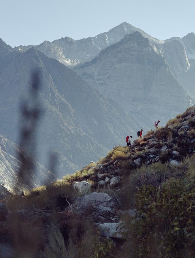 Four hikers ascend a rocky, brush-covered mountain trail with majestic peaks in the background.