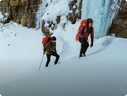 Two mountaineers ascend a snowy slope next to a large frozen waterfall.