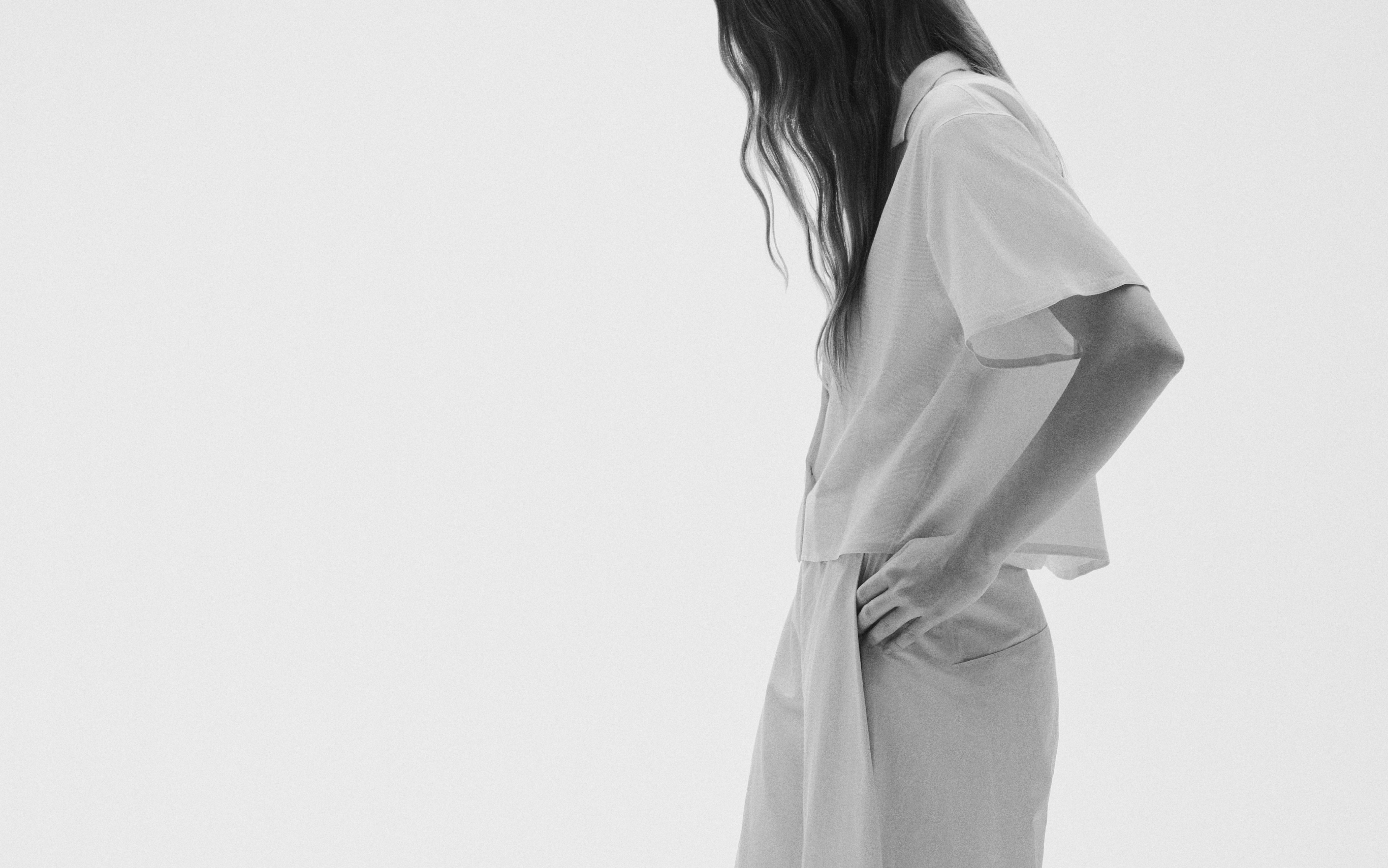 Black and white image of a person with long hair, wearing a short-sleeved shirt and pants, hand in pocket, looking down.