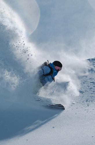 A snowboarder carves down a sunny slope, kicking up a large cloud of snow powder.