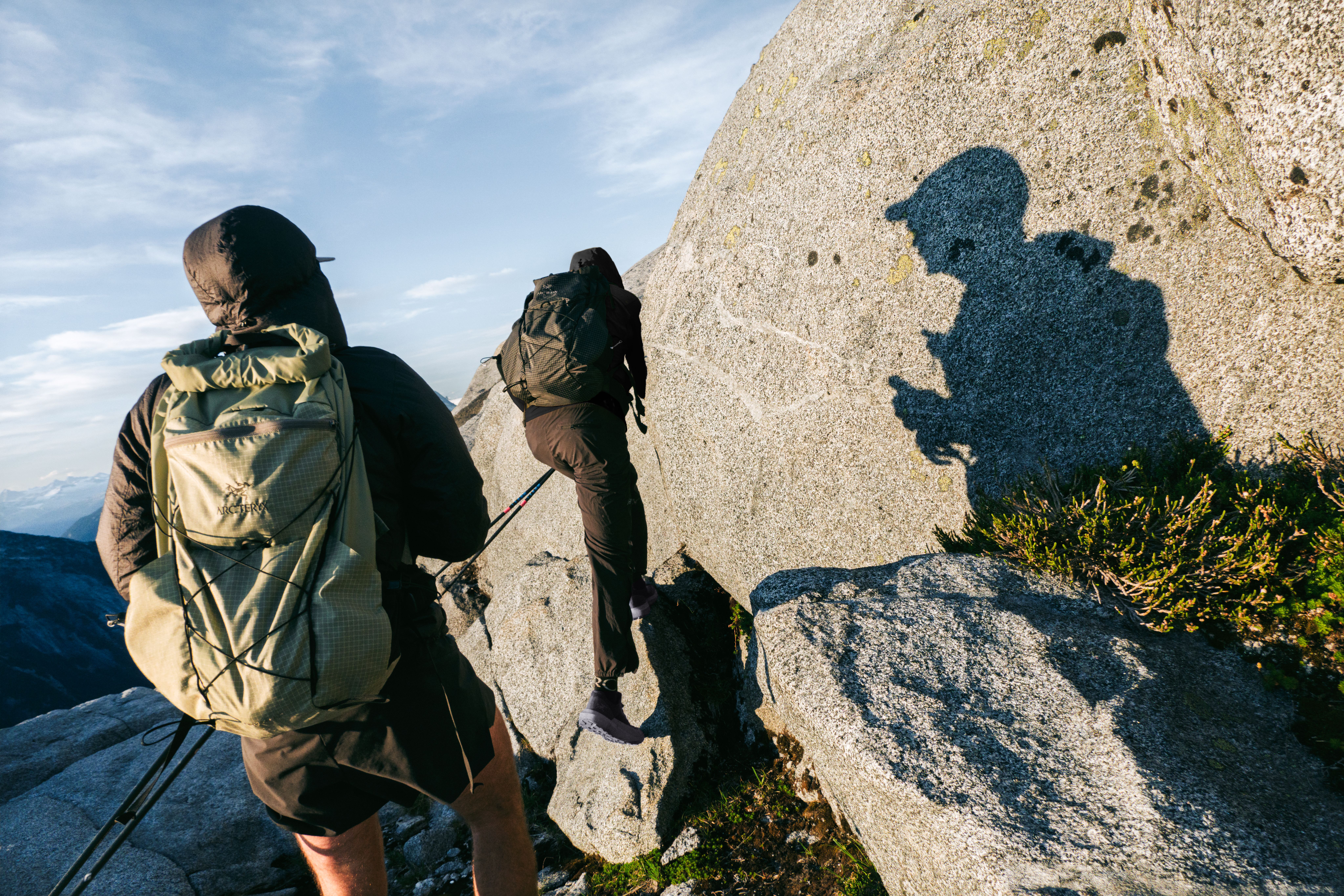 Two hikers with backpacks and trekking poles ascend a rocky mountain, one climbing a large boulder, with a person's shadow cast on a nearby rock.