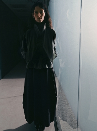 A woman in dark clothing stands in a dimly lit hallway next to a glowing frosted glass panel.