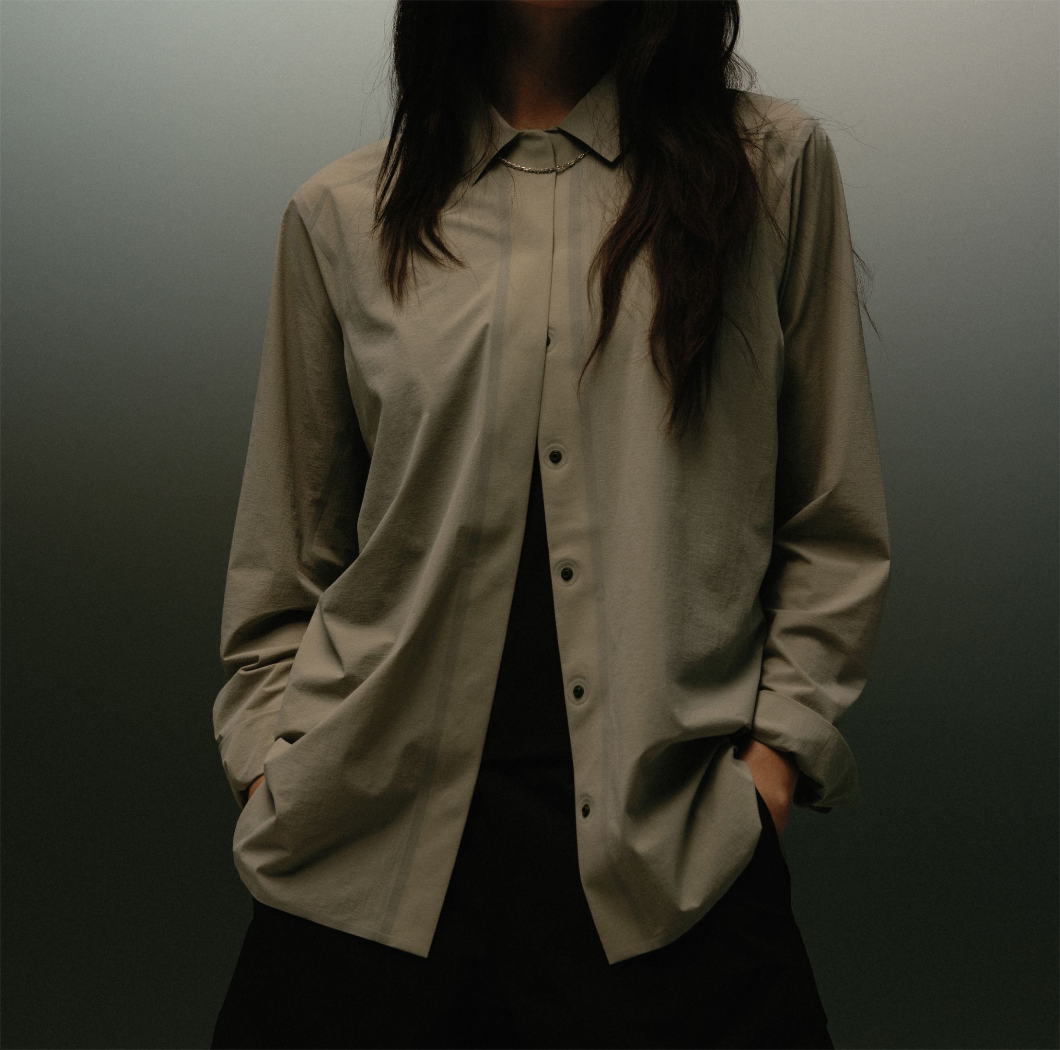 A person wearing a loose, unbuttoned beige collared shirt over black clothing, with hands in pockets.