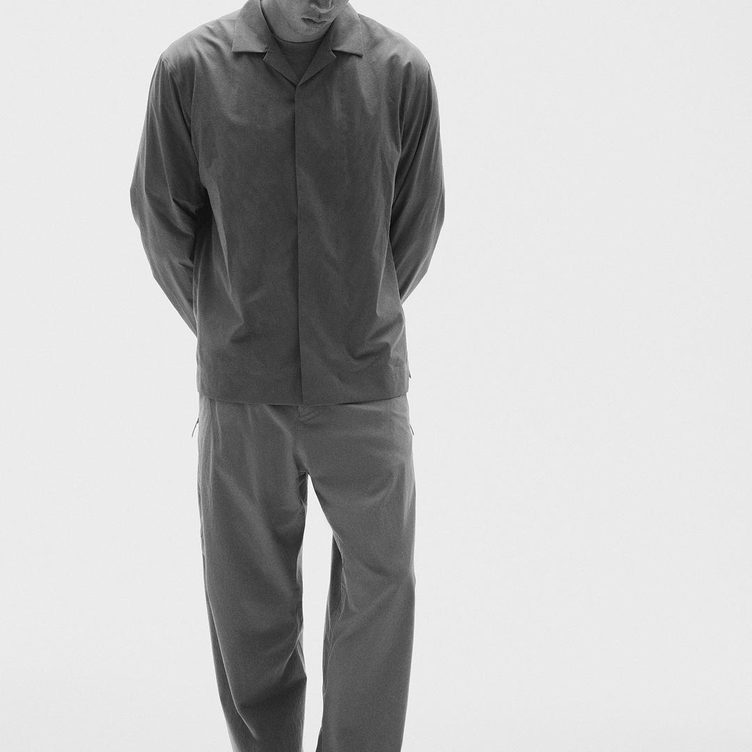 A person wearing a dark gray collared shirt and matching pants, standing with hands behind their back.