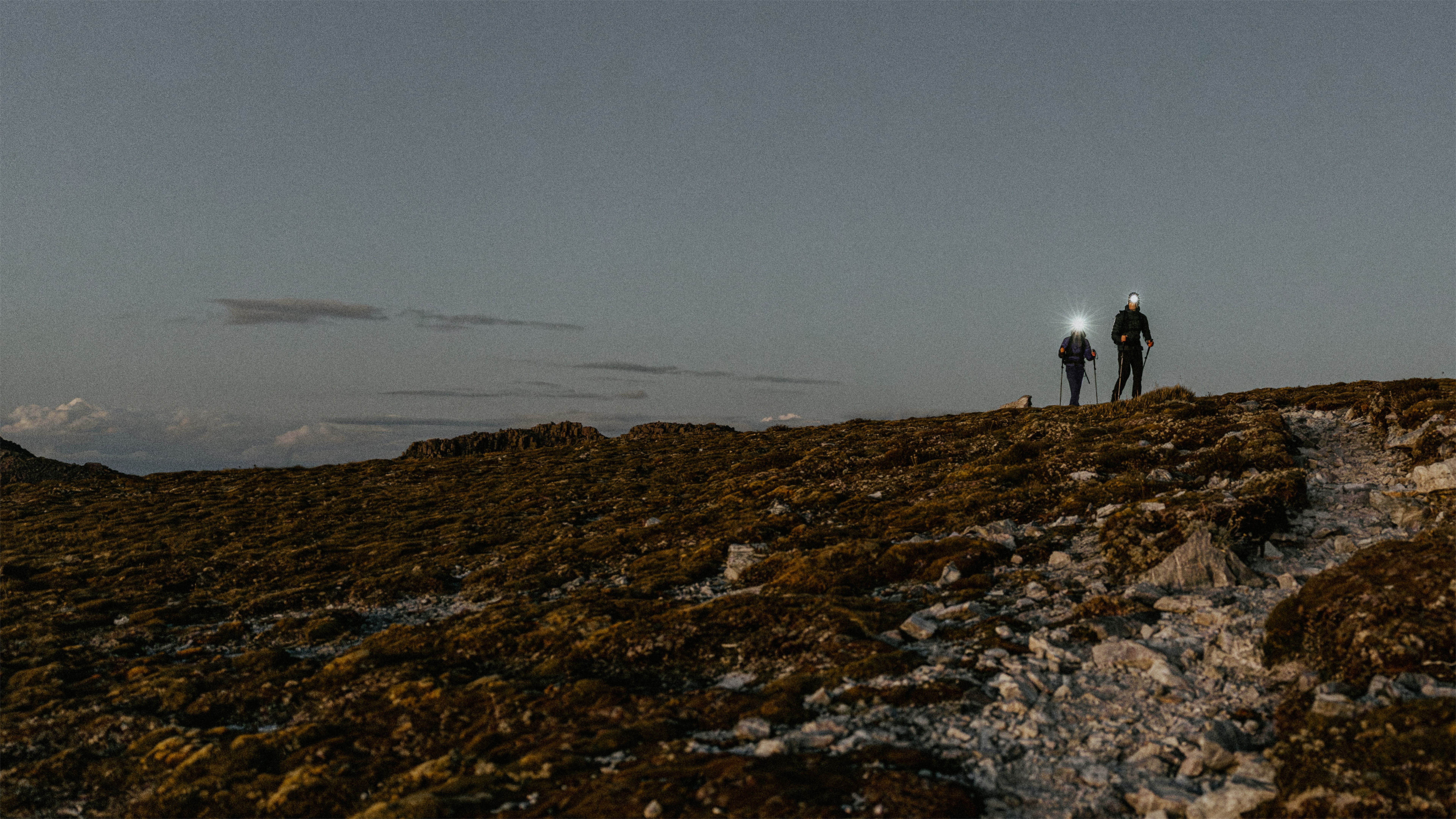 A person with a lit headlamp and backpack climbs rocky terrain in fog, with another headlamp glowing in the distance.