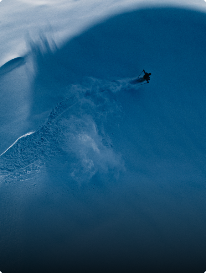 A lone skier descends a steep, shadowy blue snow slope, spraying powder.