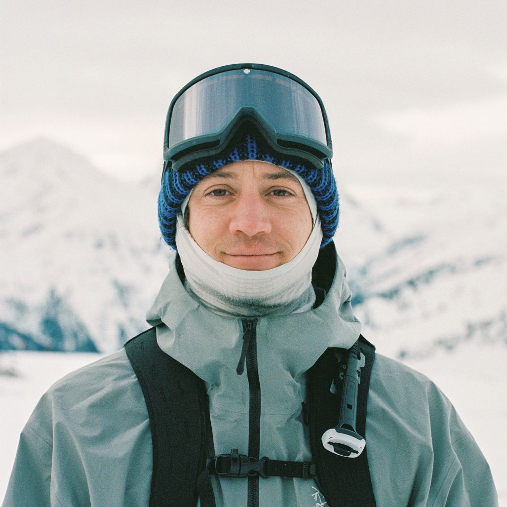 Man in ski goggles, blue hat, and neck gaiter smiling against a snowy mountain backdrop.