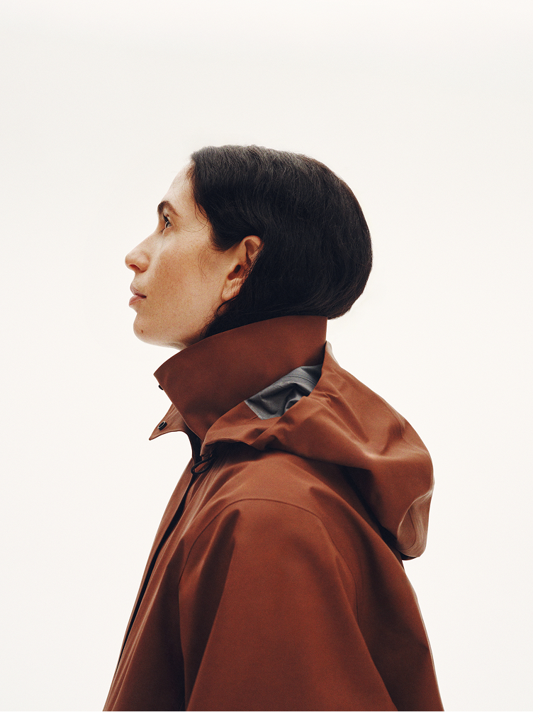 Profile of a woman in a rust-colored hooded jacket looking up.