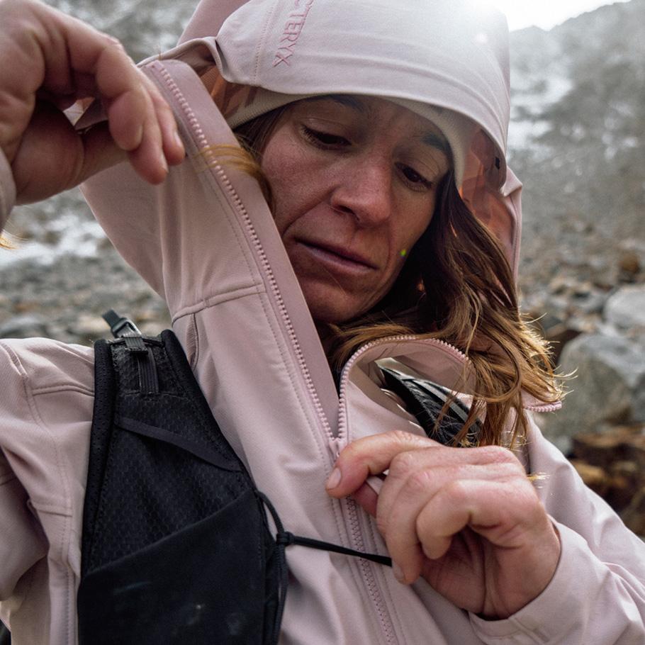 A woman in a pink Arc'teryx hooded jacket and hydration vest adjusts her jacket outdoors.
