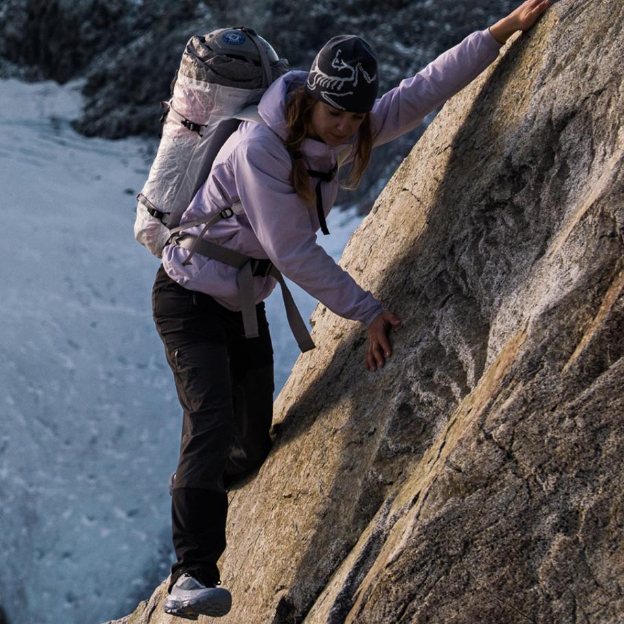 A woman in a purple jacket and backpack scrambles up a rock face with snow in the background.