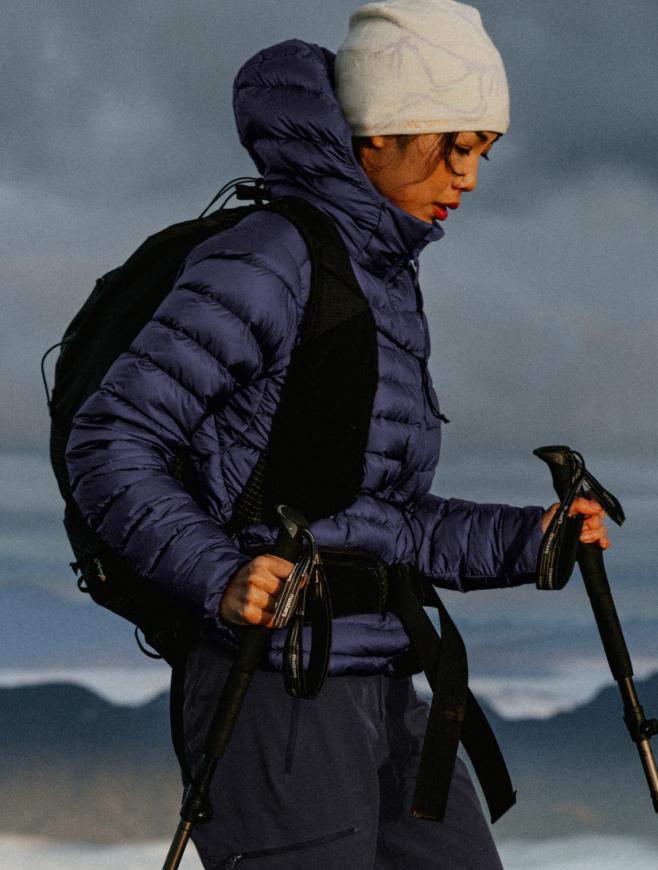 A woman in a purple puffer jacket, white beanie, and backpack holds trekking poles while hiking against a cloudy mountain background.