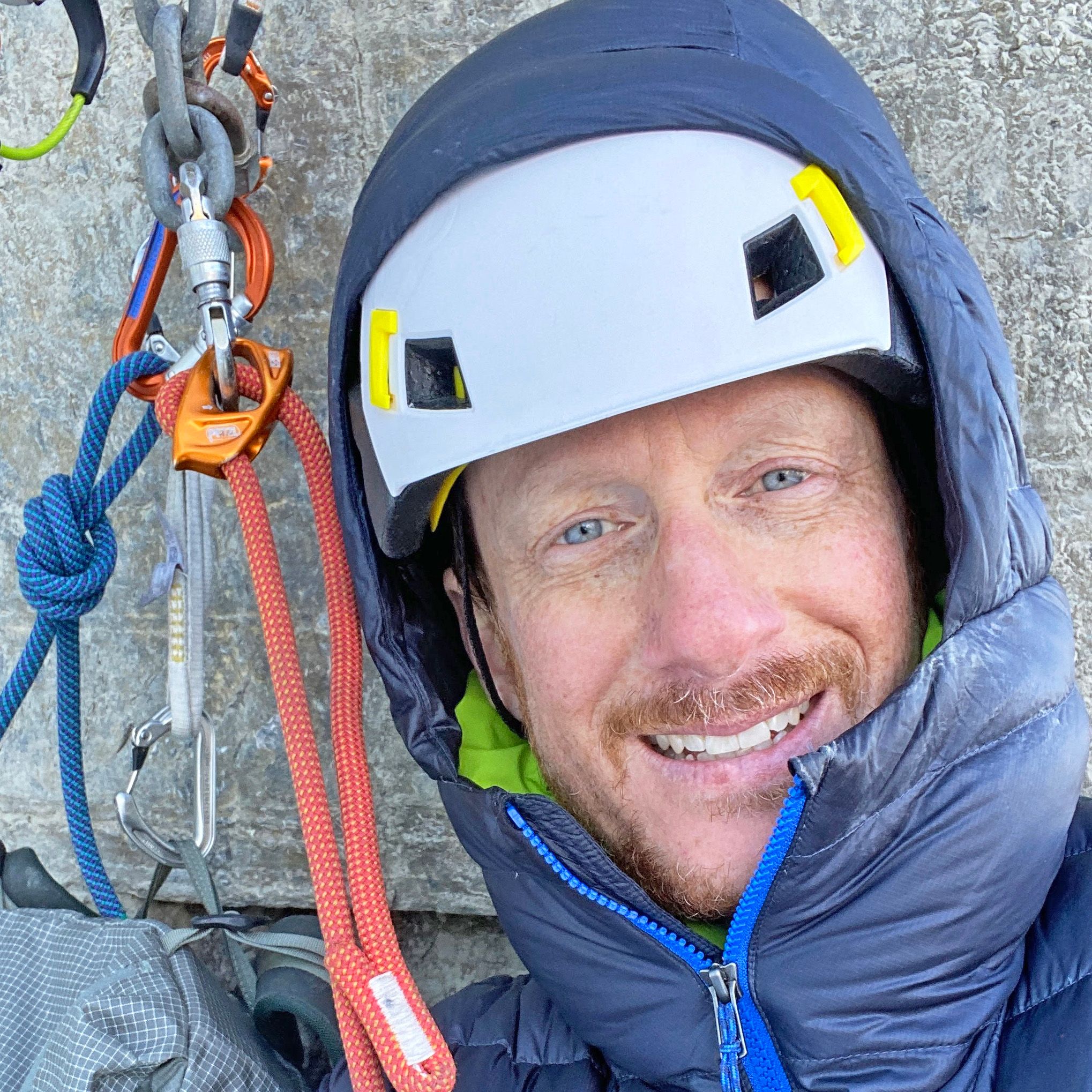 Smiling climber in a helmet and hooded jacket with climbing gear.
